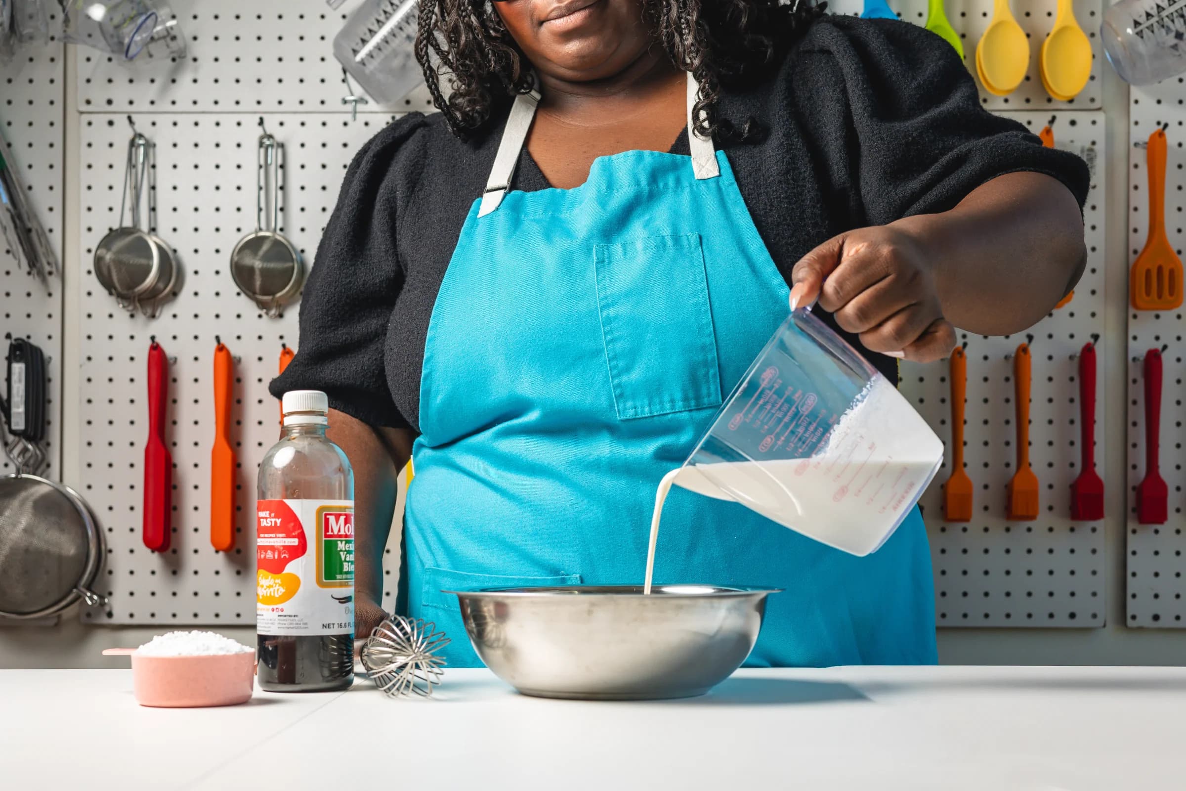 A cook in a turquoise apron pours ingredients into a mixing bowl
